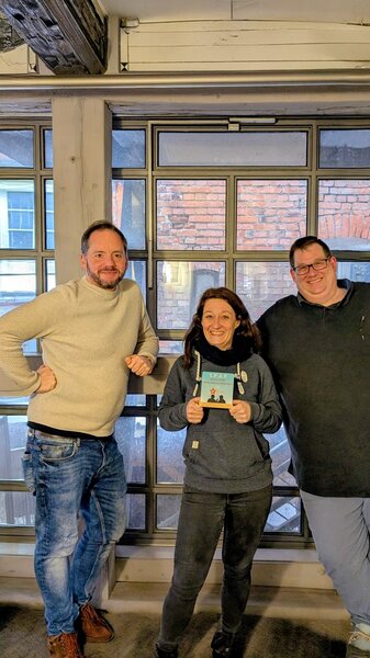 Pjär, Ina Hoinkis und Sascha stehen gemeinsam vor einem Fenster; Ina hält die SPÄR-Danksagung in den Händen.