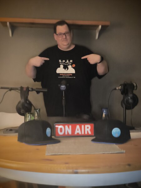 Sascha steht im SPÄR-Studio zwischen Mikrofonen und On-Air-Schild und zeigt auf sein SPÄR-Shirt.